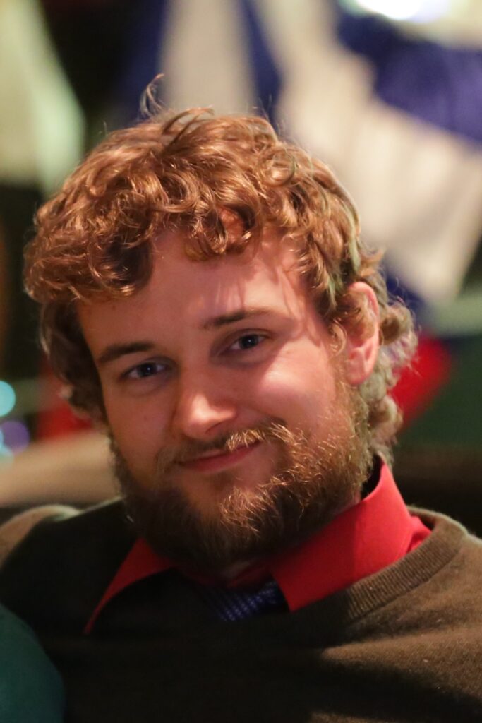 A white man with medium length brown curly hair, a beard, in semi-casual clothes and a sweater.